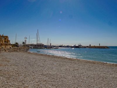 Une large plage de galets s'étend vers une marina remplie de yachts, bordée de bâtiments côtiers traditionnels sous un ciel bleu clair.