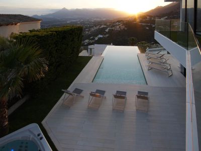 Vaste piscine à débordement avec terrasse claire, chaises longues et jacuzzi séparé, offrant une vue panoramique sur un coucher de soleil montagneux.
