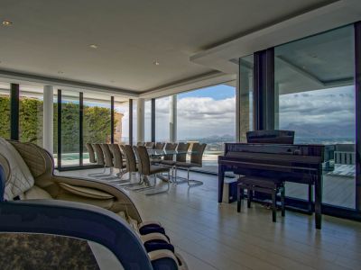 Vaste salon-salle à manger avec table en verre pour dix, piano à queue et fauteuil de massage. Grandes baies vitrées offrant des vues panoramiques sur les montagnes et le paysage.
