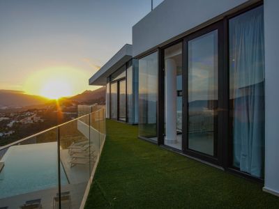 Vaste terrasse avec piscine à débordement, pont en bois et balustrade en verre, offrant des vues panoramiques sur le coucher de soleil montagneux.