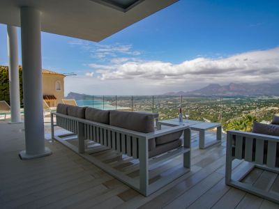 Vaste terrasse couverte avec plancher clair et mobilier lounge blanc moderne, offrant une vue panoramique sur la mer, la côte et les montagnes via une balustrade vitrée.