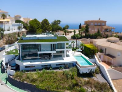 Vue aérienne de la villa moderne blanche au design épuré avec toit végétalisé, piscine à débordement et jacuzzi séparé, offrant une vue sur la mer.