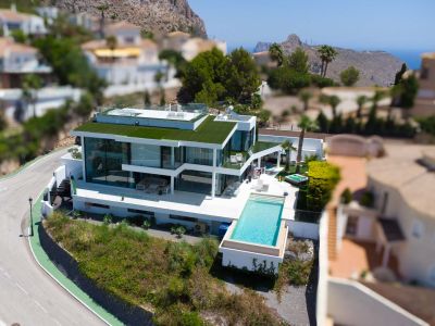 Vue aérienne d'une villa blanche contemporaine avec piscine à débordement, façades vitrées, toit végétalisé et jacuzzi, offrant des vues sur la montagne et la mer.