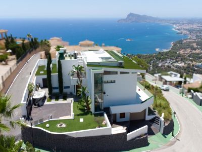 Vue aérienne d'une villa blanche moderne à plusieurs niveaux, avec toits plats végétalisés et ascenseur extérieur en verre. Elle dispose d'une allée privée et de vues panoramiques sur la mer et la côte.