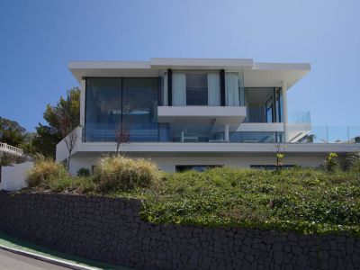 Vue extérieure d'une villa moderne à trois niveaux avec de vastes façades vitrées et balustrades en verre, surplombant un mur de pierre et la végétation le long d'une route courbée.