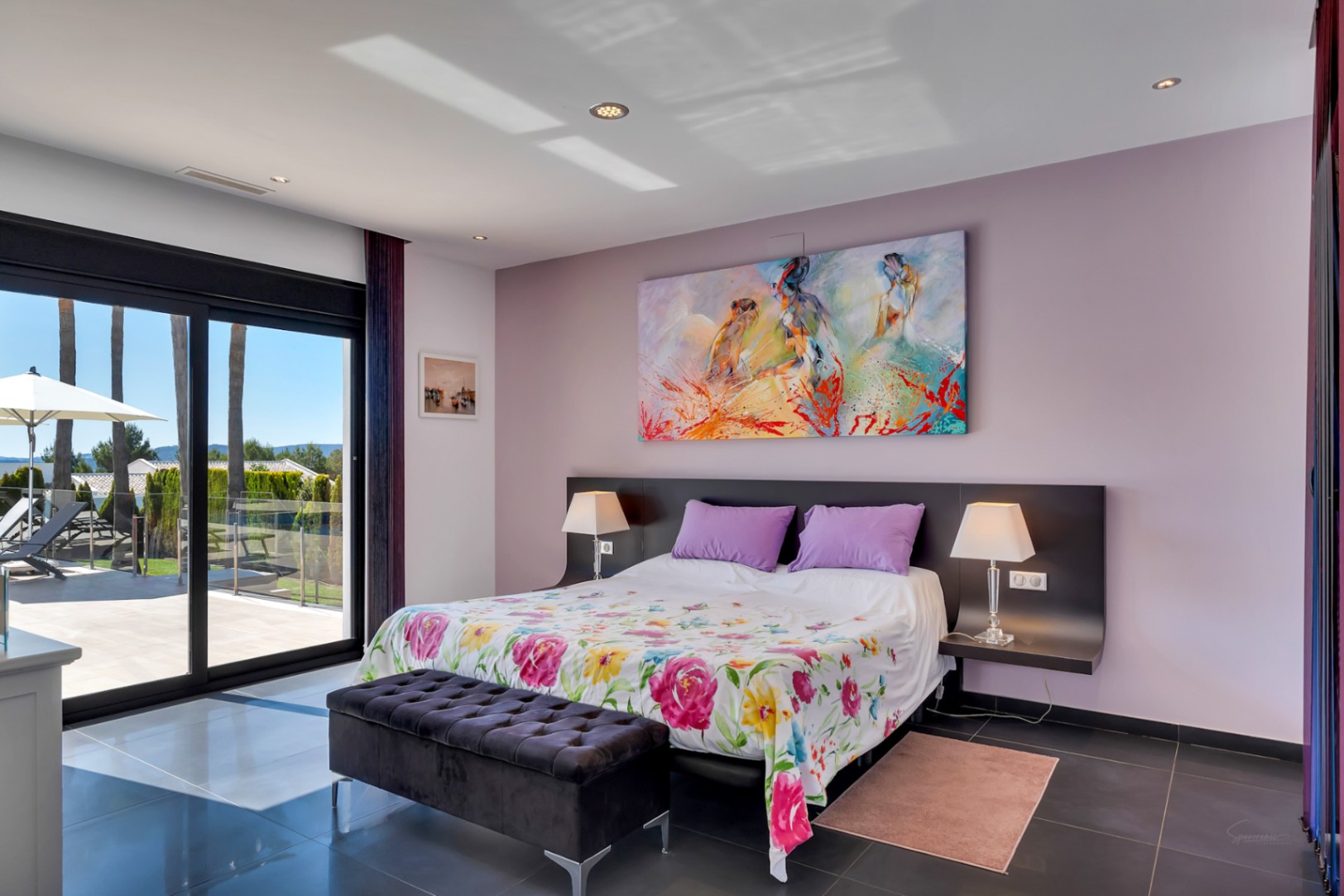 Chambre à coucher avec accès direct à la terrasse par de grandes portes coulissantes en verre, sol carrelé foncé, lit orné d'une couette florale et tableau abstrait coloré. Vues sur les palmiers extérieurs.