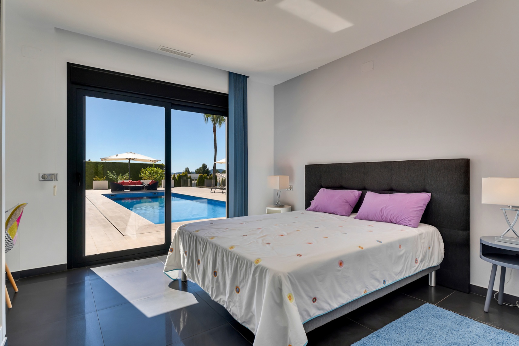 Chambre avec carrelage foncé et accès direct à la piscine extérieure, comprenant un espace détente avec canapé rouge et parasol sous un ciel bleu clair.