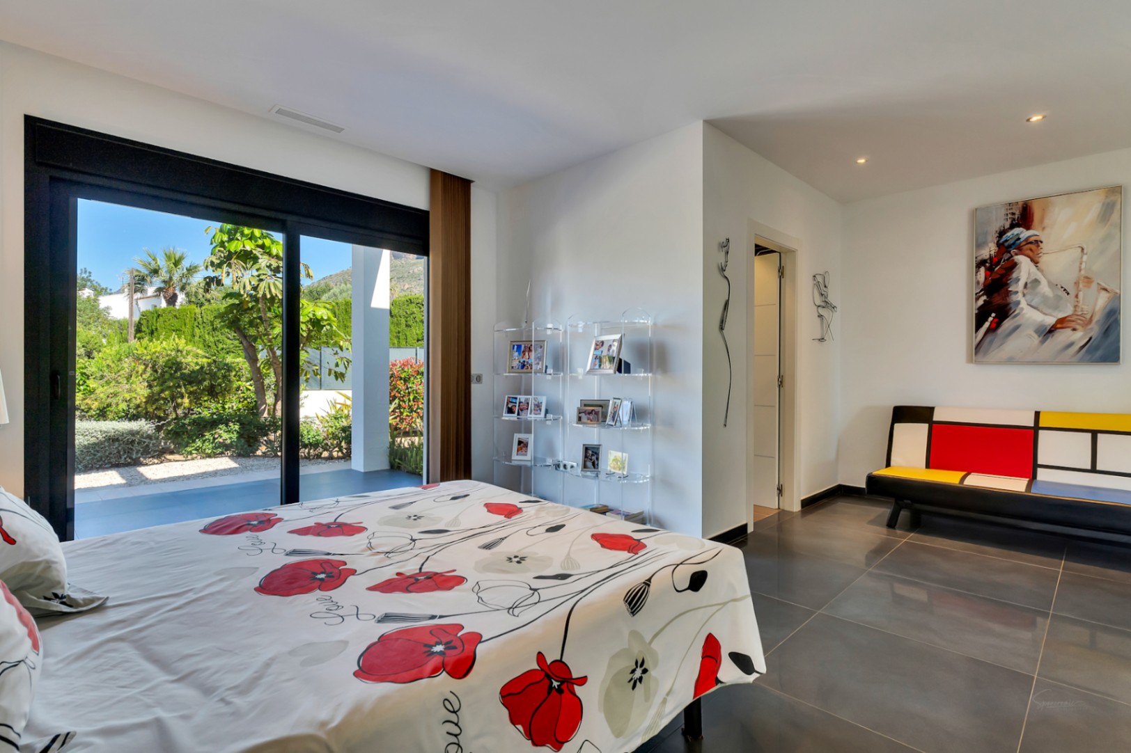 Chambre avec de grandes portes coulissantes vitrées s'ouvrant sur un jardin méditerranéen luxuriant, sol en carrelage foncé et mobilier moderne.