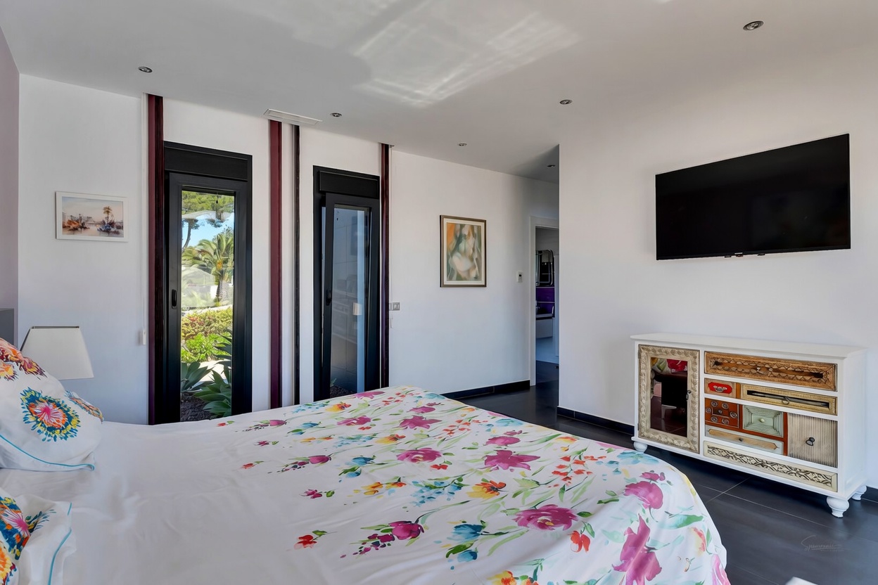 Chambre lumineuse avec lit à motifs floraux, accès direct au jardin par des portes vitrées et une commode unique à tiroirs variés.