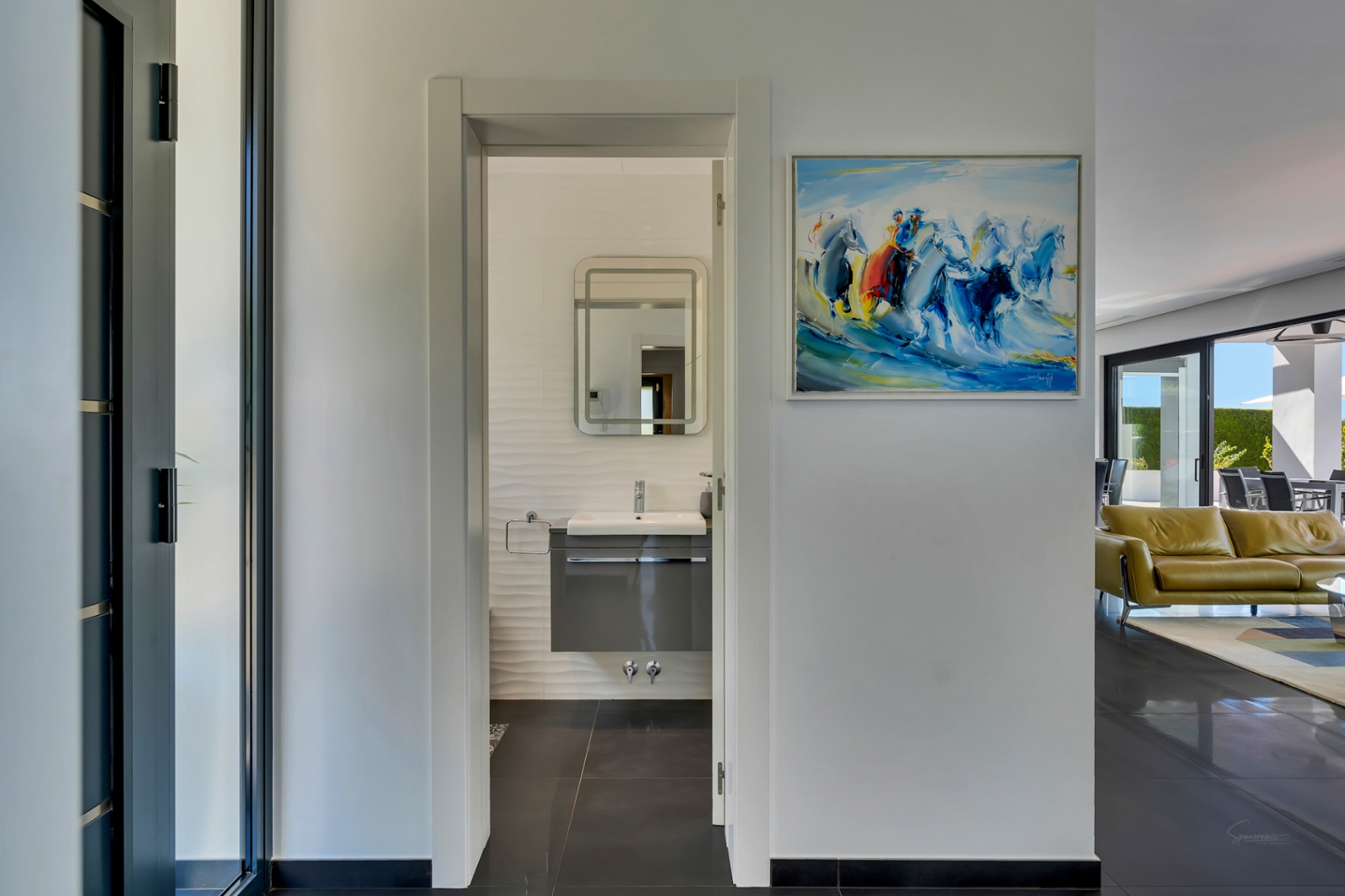 Couloir lumineux vers une salle de bain moderne avec meuble vasque suspendu, murs texturés et miroir éclairé. Un tableau abstrait et un aperçu du salon avec terrasse sont visibles.