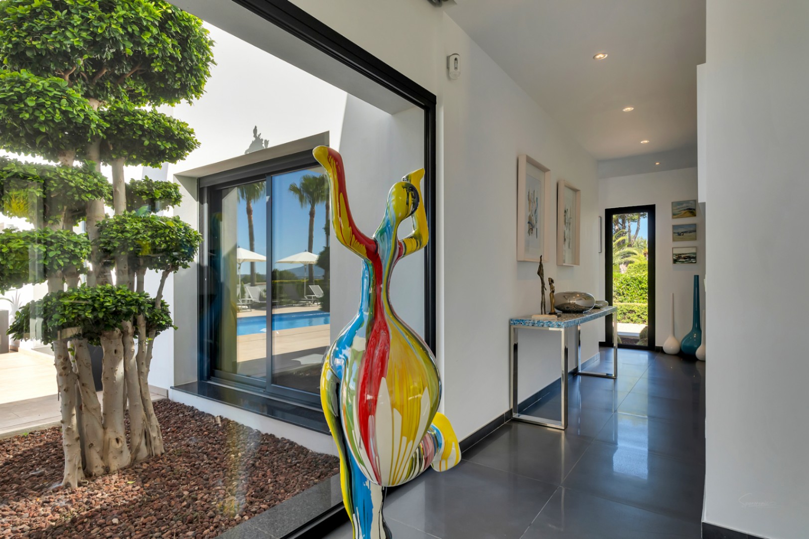 Couloir moderne aux carreaux sombres, orné d'une sculpture abstraite colorée et offrant une vue sur la terrasse piscine et un arbre décoratif intérieur.