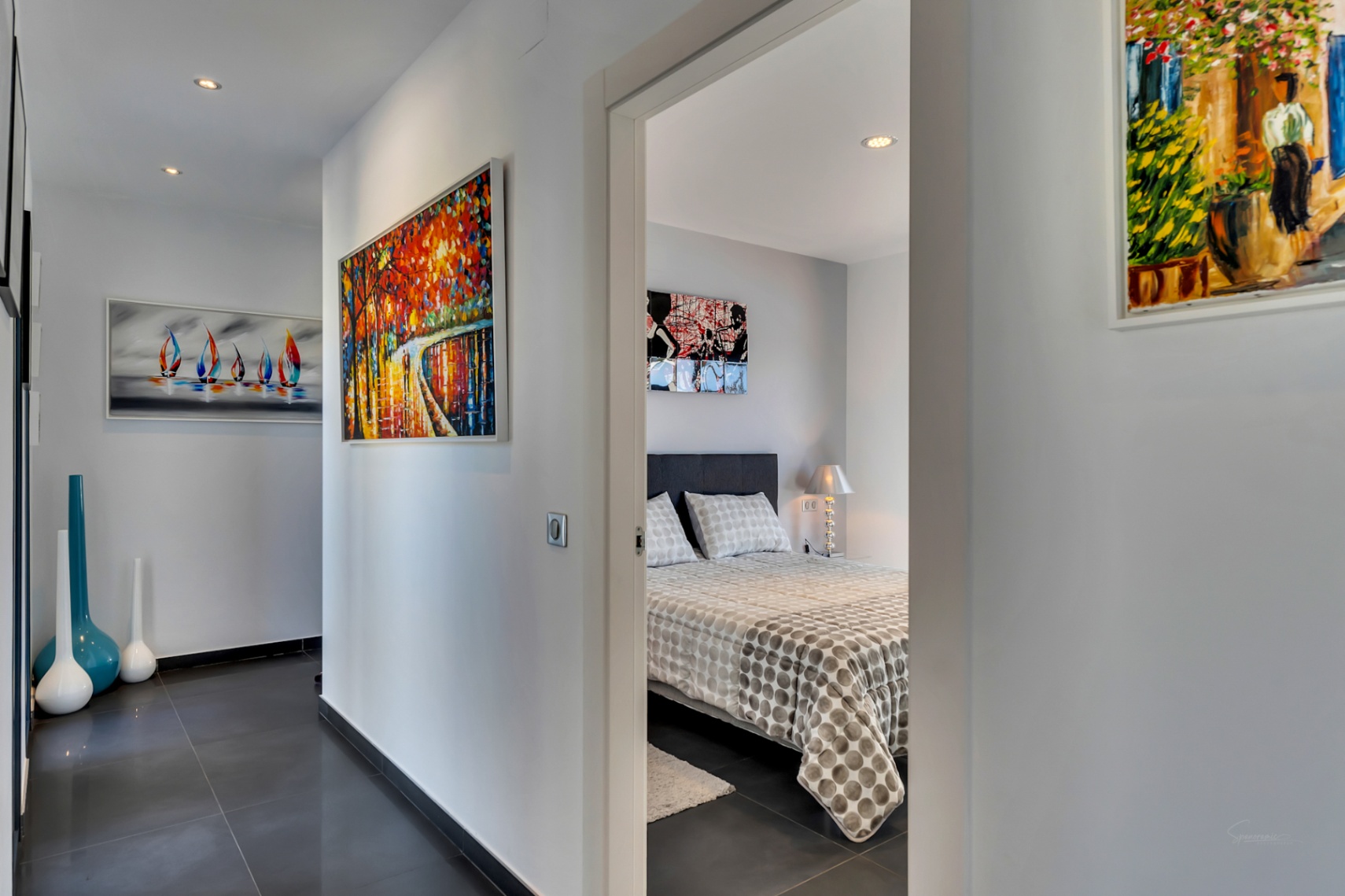 Couloir moderne avec carrelage gris foncé, éclairage encastré et art abstrait coloré, menant à une chambre avec lit à motifs et lampe de chevet.
