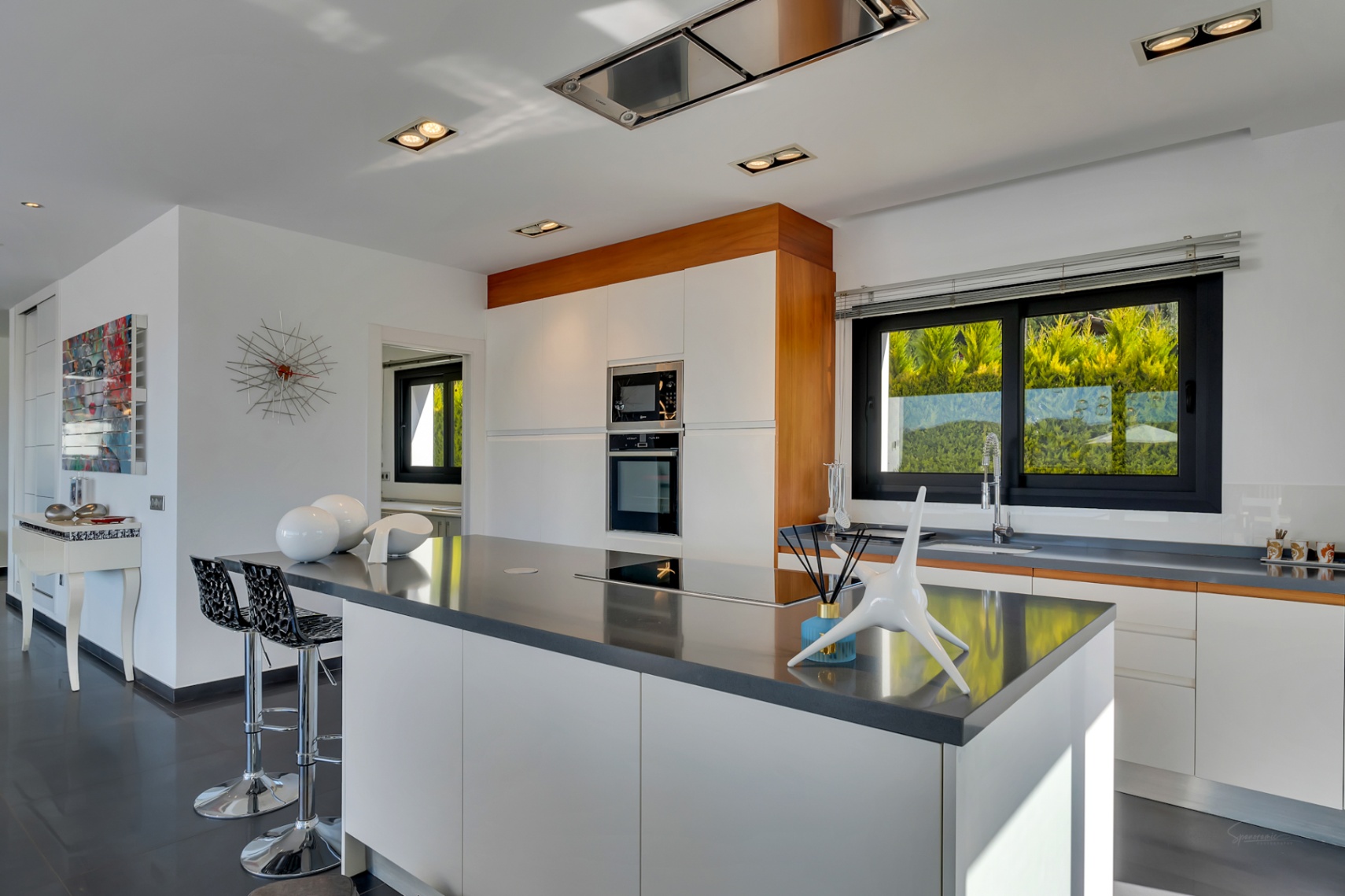 Cuisine moderne avec grand îlot central à plaque intégrée, meubles blancs sans poignées et électroménager encastré. Grande fenêtre offrant lumière naturelle et vue sur la verdure.