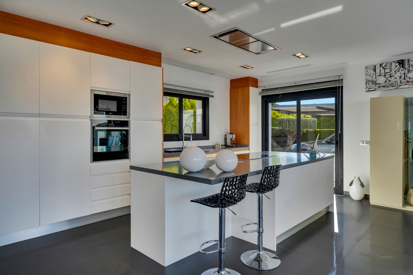 Cuisine moderne avec grand îlot central et sièges, électroménager intégré, armoires blanches et accès direct à l'extérieur par porte coulissante vitrée.