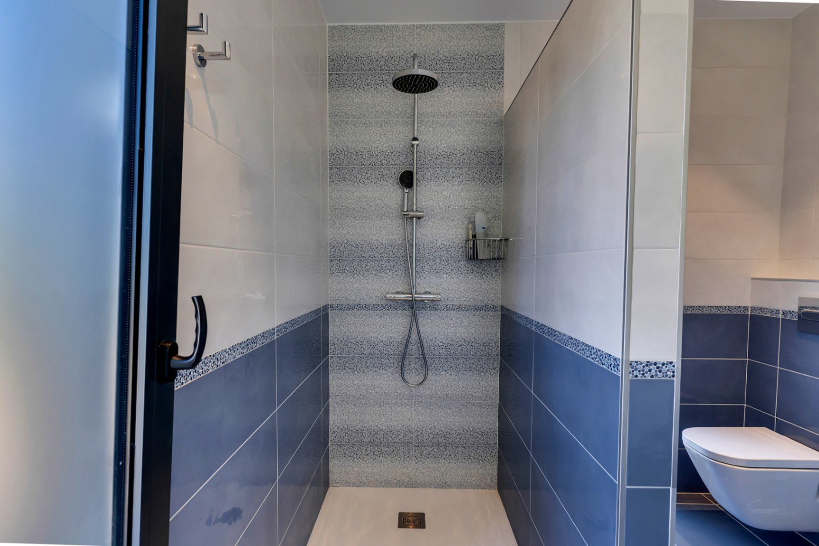 Douche à l'italienne carrelée de bleu et blanc avec une bande décorative en mosaïque. Comprend une pomme de douche pluie et une douchette, séparée par des parois vitrées.