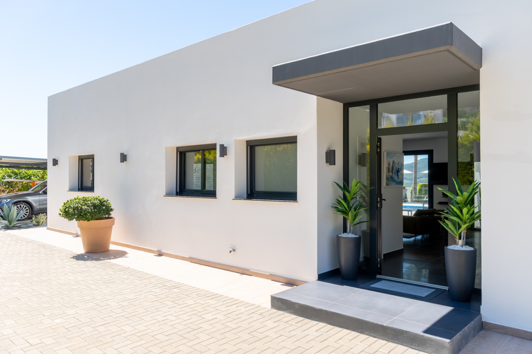 Entrée moderne d'une villa blanche avec porte vitrée encadrée de noir et auvent gris. Extérieur pavé, fenêtres à volets et parking couvert visible.