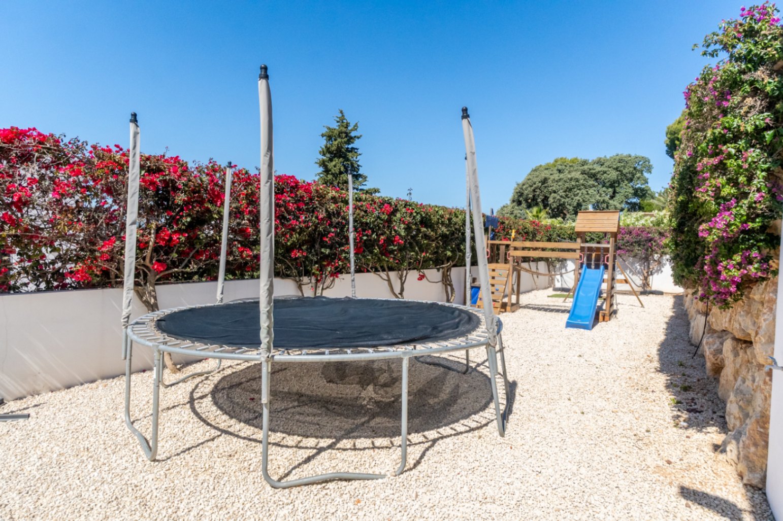 Espace de jeux extérieur avec trampoline et structure en bois avec toboggan bleu sur gravier. Des bougainvilliers luxuriants et un mur de pierre avec des vignes violettes encadrent la zone sous un ciel clair.