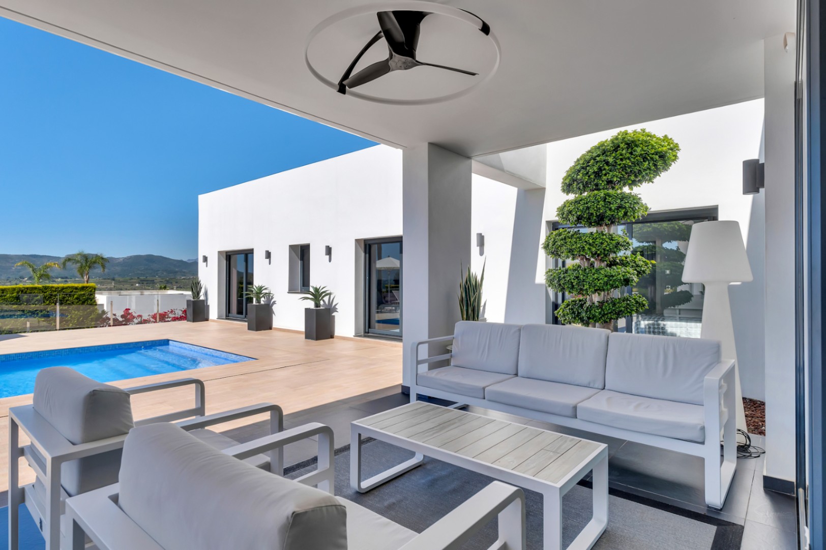 Espace lounge extérieur couvert avec mobilier blanc contemporain, ventilateur de plafond et accès direct à la piscine. Architecture minimaliste et vue sur les montagnes.