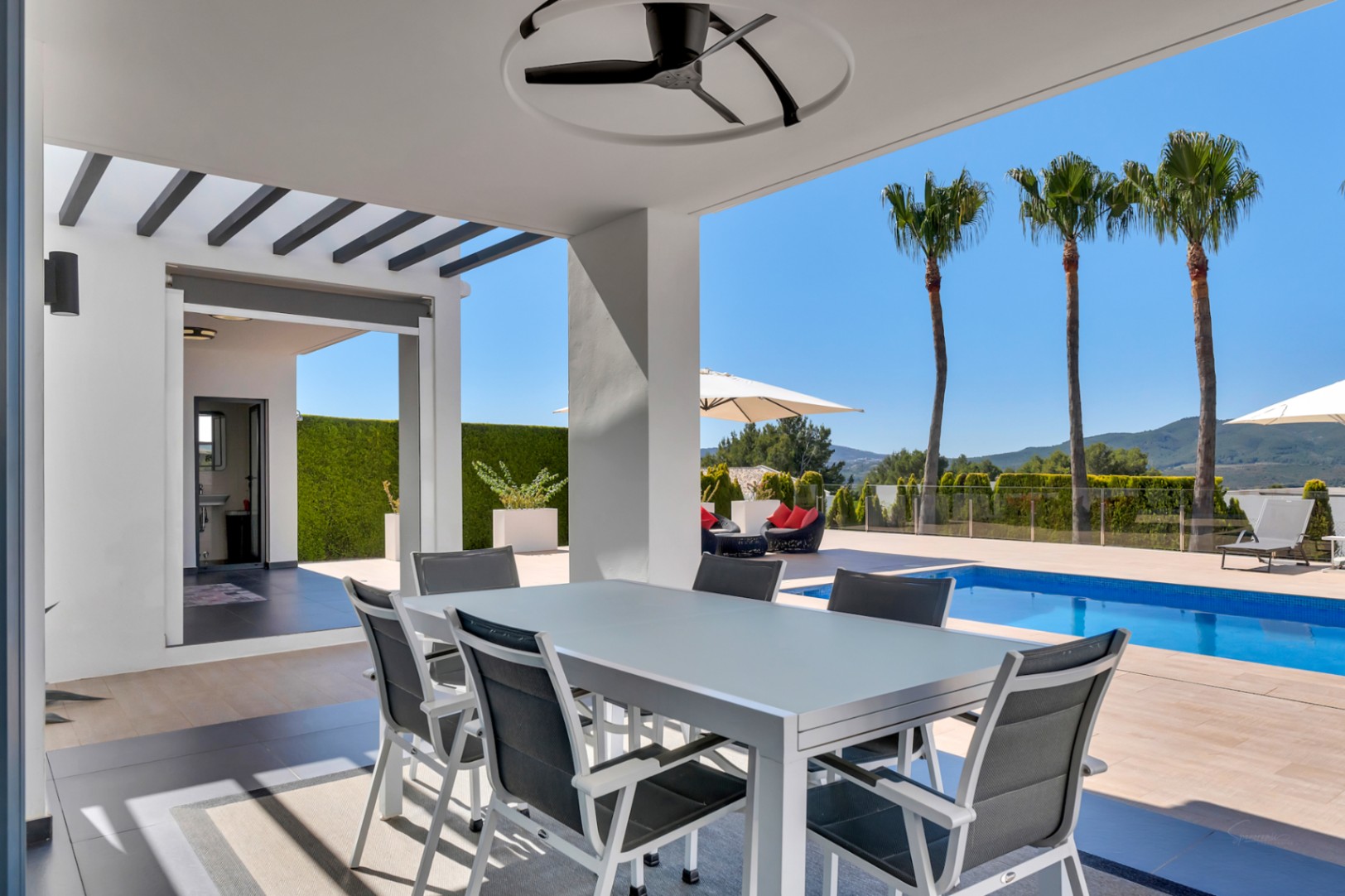 Espace repas extérieur couvert avec table et chaises modernes, ventilateur de plafond, vue sur la piscine, les palmiers et les montagnes. Une salle de bain extérieure est visible.