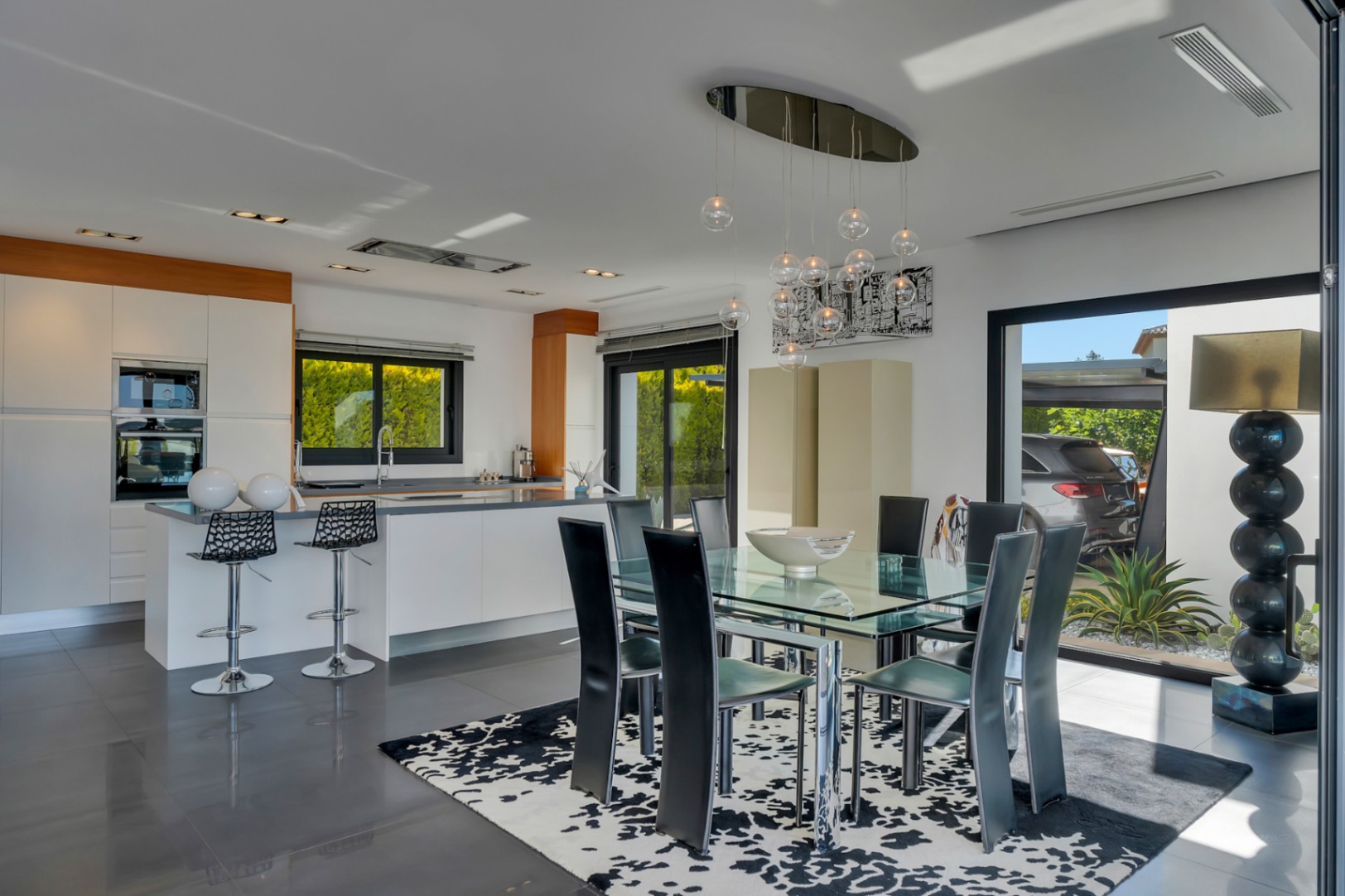 Espace repas intégré à une cuisine moderne blanche, avec table en verre, chaises noires et lustre contemporain. De grandes portes coulissantes mènent à l'extérieur.