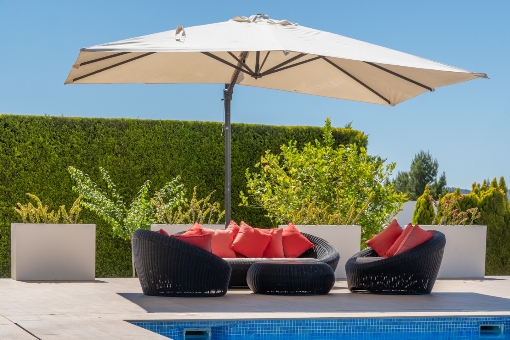 Espace salon extérieur avec mobilier en rotin noir et coussins rouges vifs, ombragé par un grand parasol, à côté de la piscine carrelée.