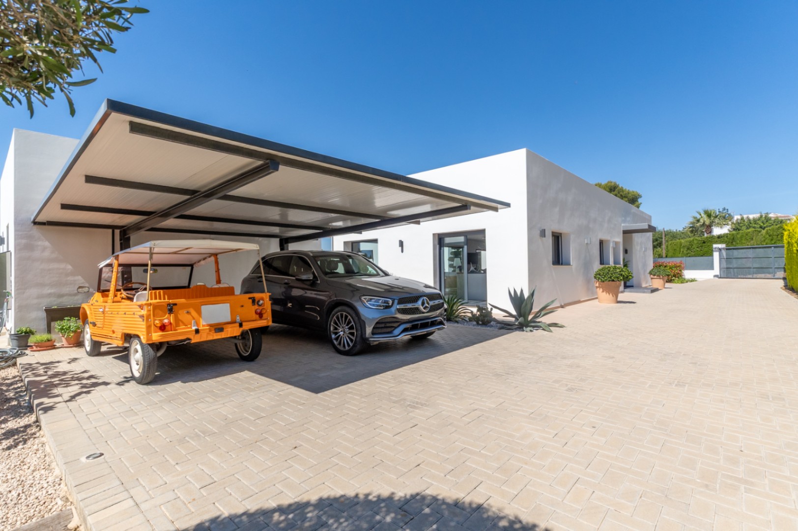 Extérieur de villa moderne avec double abri de voiture couvert à structure sombre, abritant un SUV Mercedes et une voiture orange décapotable, sur une allée pavée.