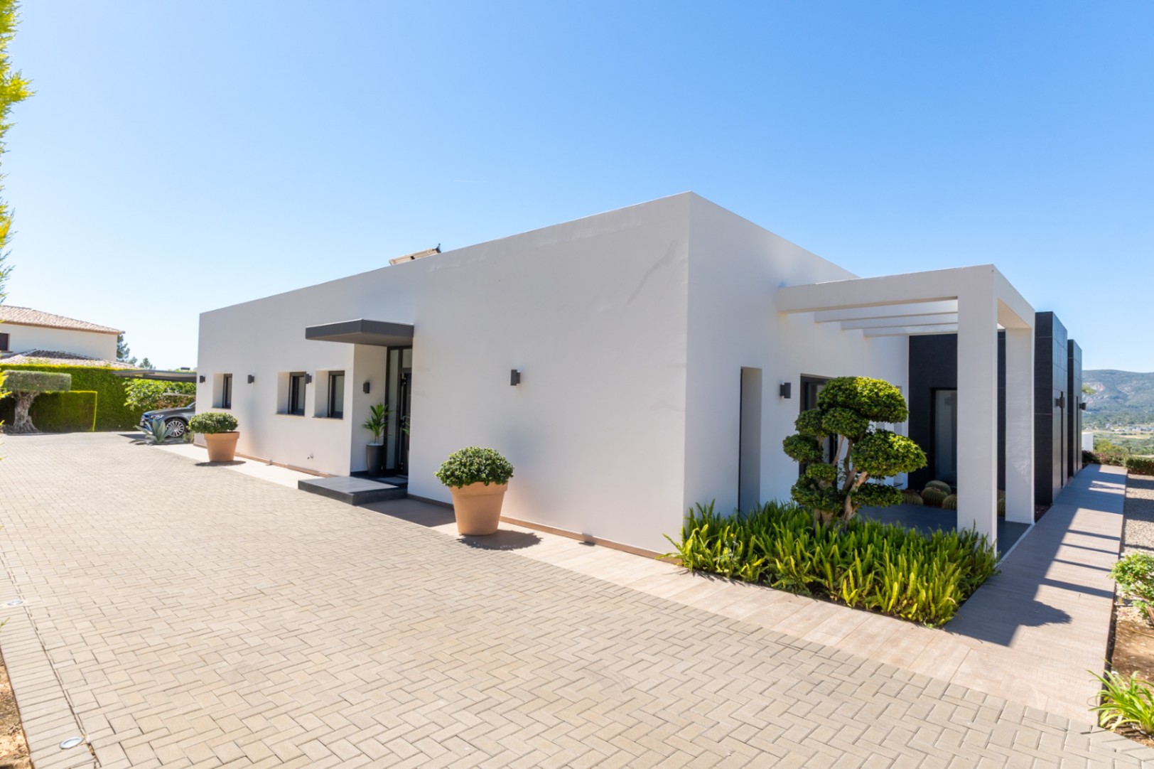 Extérieur de villa moderne avec façade blanche épurée, allée pavée et aménagement paysager minimaliste. Arbre sculpté, pergola et collines lointaines.