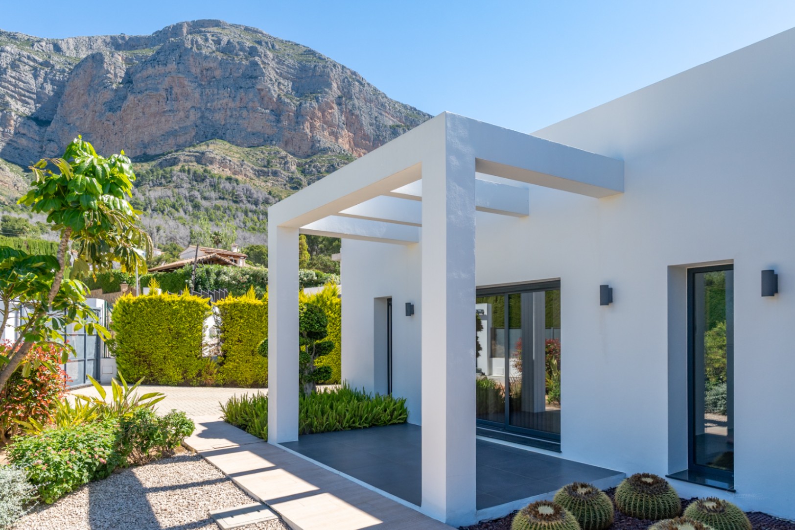 Extérieur moderne de la villa avec pergola blanche, jardin méditerranéen et vues dégagées sur l'imposante montagne du Montgó.