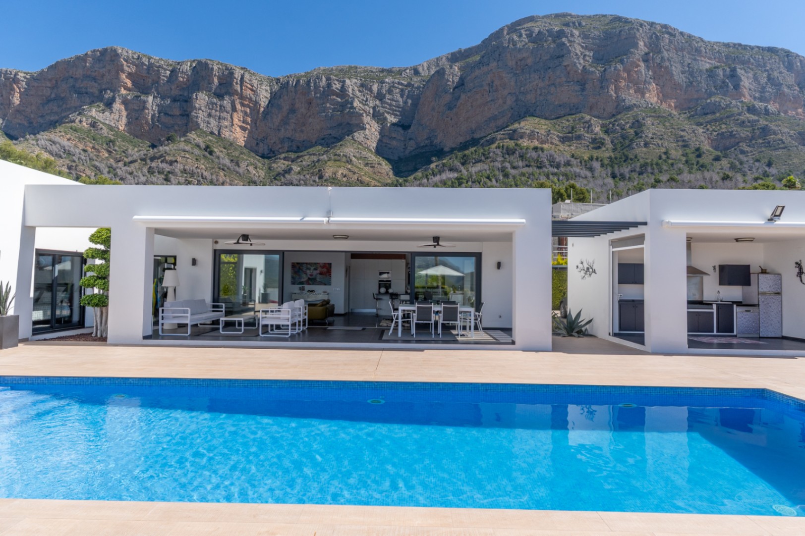 La villa moderne offre une piscine bleue lumineuse, une terrasse couverte avec salon et salle à manger extérieurs, et une cuisine d'été intégrée, face au Montgó.