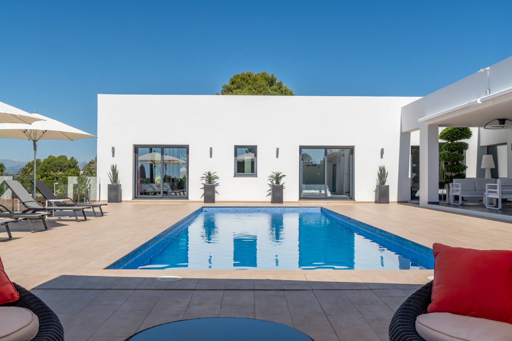 Piscine rectangulaire en mosaïque bleue sur une terrasse carrelée, devant une façade blanche moderne avec de grandes portes coulissantes et un salon extérieur couvert.