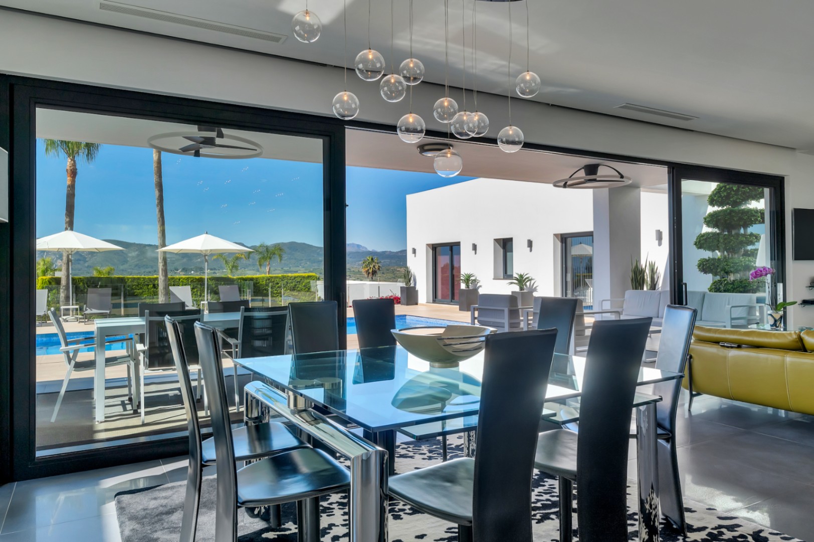Salle à manger moderne avec table en verre et chaises noires, s'ouvrant sur une terrasse piscine avec transats, palmiers et vue sur les montagnes.