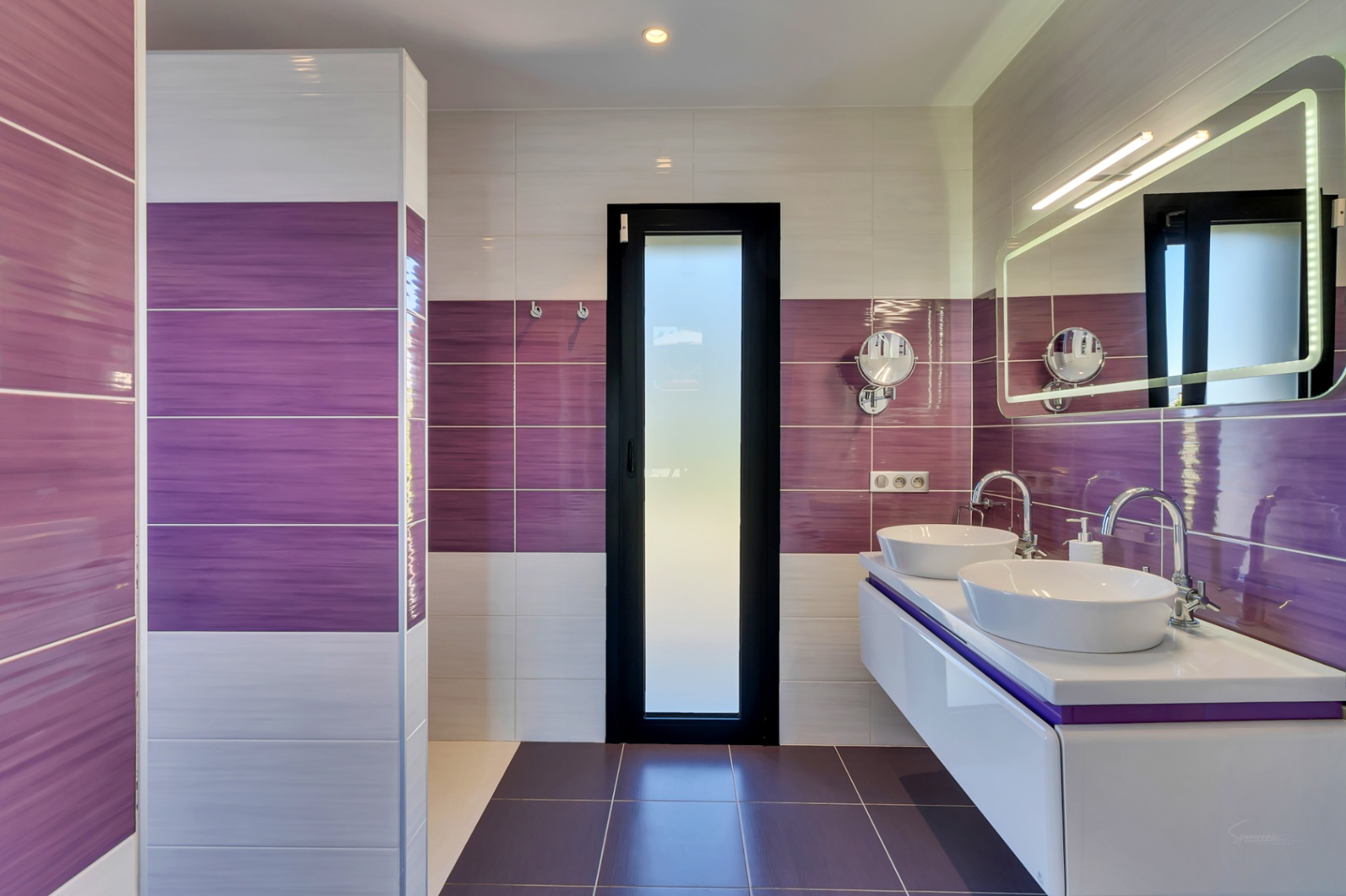 Salle de bain contemporaine avec carrelage mural violet et blanc, double vasque flottante avec lavabos ovales et miroir éclairé. Une porte en verre dépoli assure l'intimité.