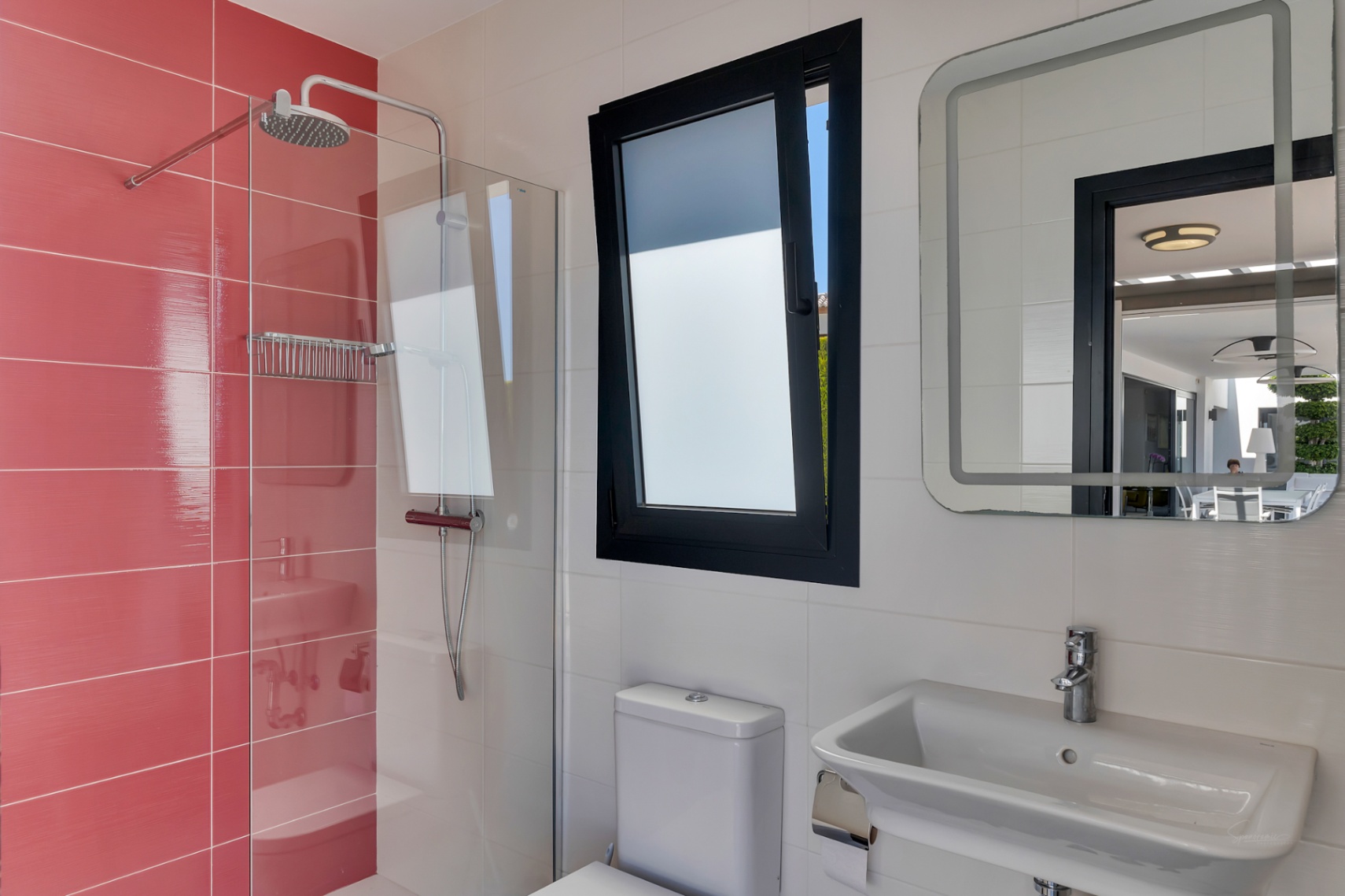 Salle de bain fonctionnelle avec douche à l'italienne carrelée en rouge, fenêtre oscillo-battante et miroir lumineux reflétant la terrasse extérieure.