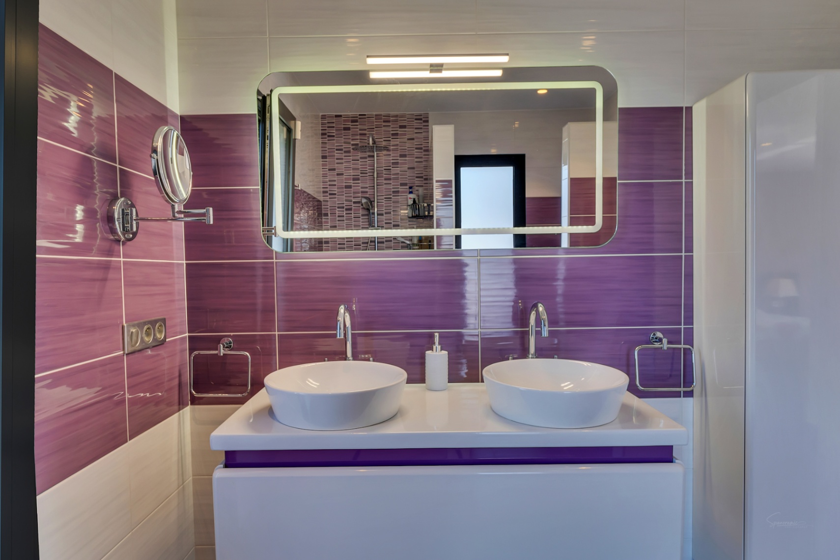 Salle de bain moderne avec double vasque, carrelage mural violet horizontal et miroir lumineux reflétant la douche en mosaïque.