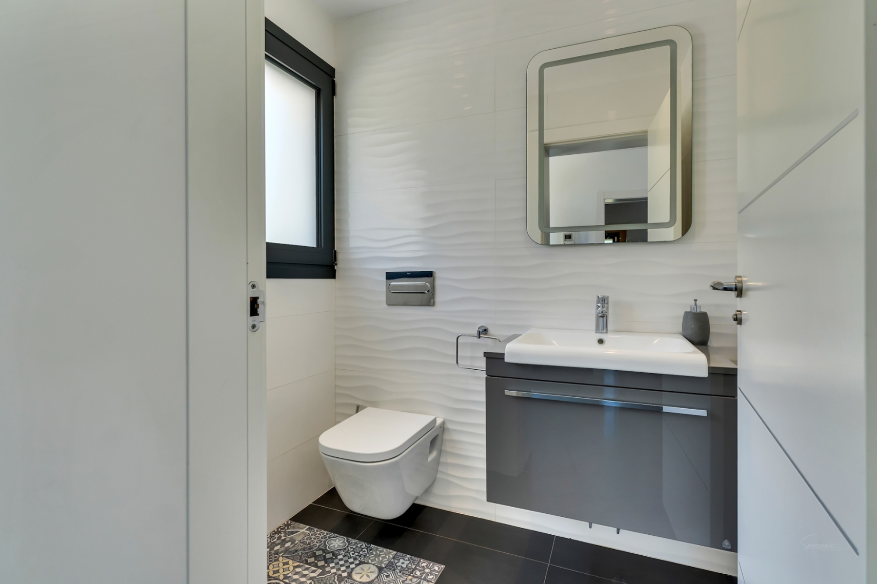 Salle de bain moderne avec toilettes suspendues, meuble vasque gris flottant et carrelage mural blanc texturé. Un miroir éclairé et des carreaux de sol décoratifs complètent l'espace.
