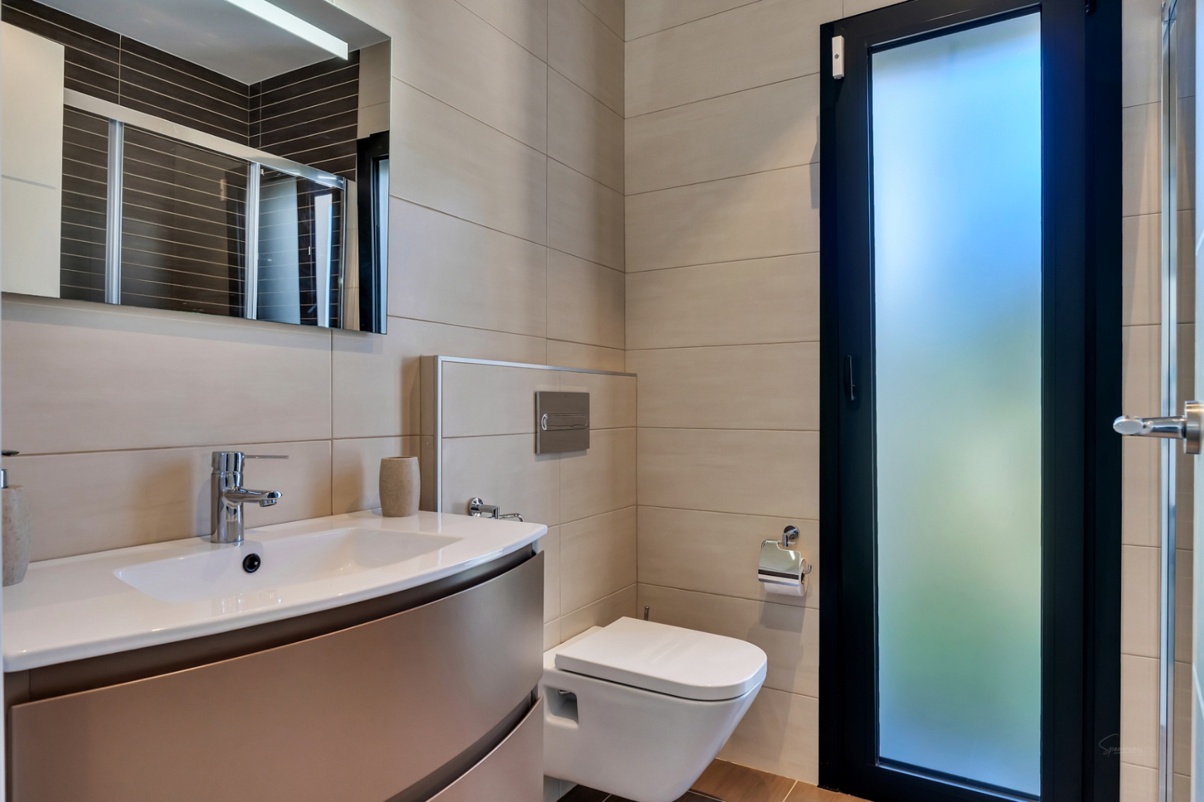 Salle de bain moderne avec WC suspendu, meuble vasque intégré, robinetterie chromée et carrelage mural neutre. Porte vitrée dépolie pour une lumière naturelle.