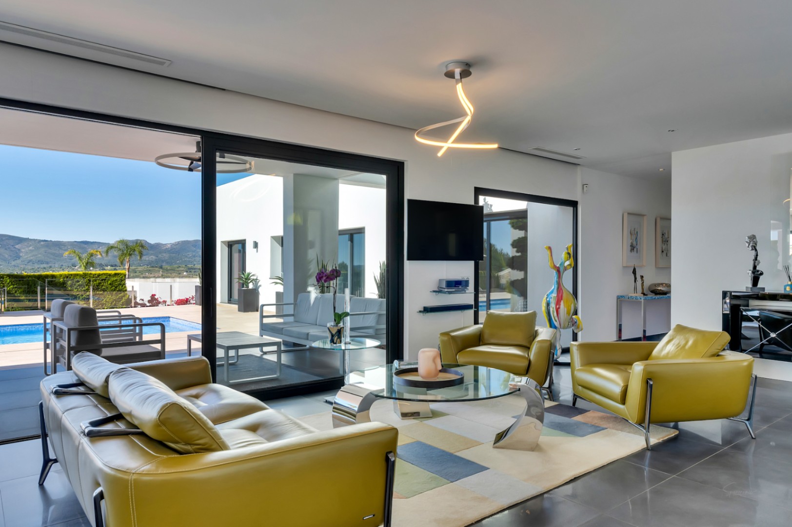 Salon lumineux avec canapé et fauteuils en cuir vert olive, table basse en verre et accès direct à la terrasse, piscine et vue sur la montagne.