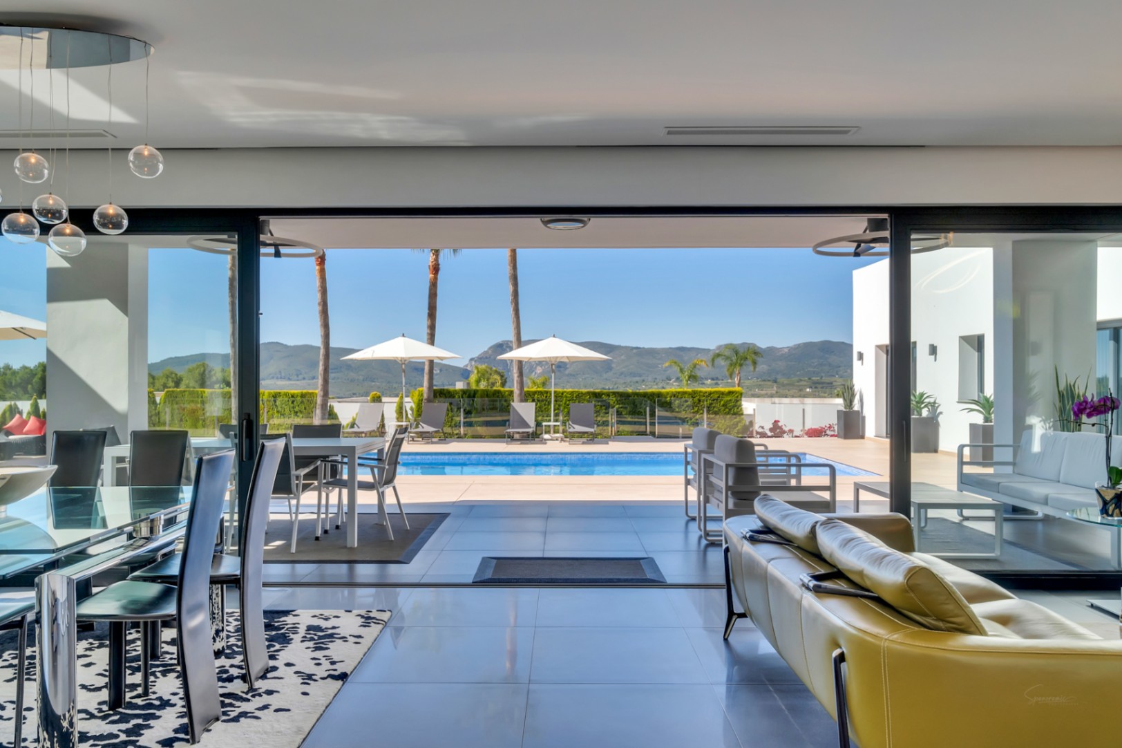 Salon-salle à manger ouvert avec table en verre, suspensions modernes et canapé jaune, s'ouvrant sur la terrasse de la piscine avec transats et vues lointaines sur les montagnes.