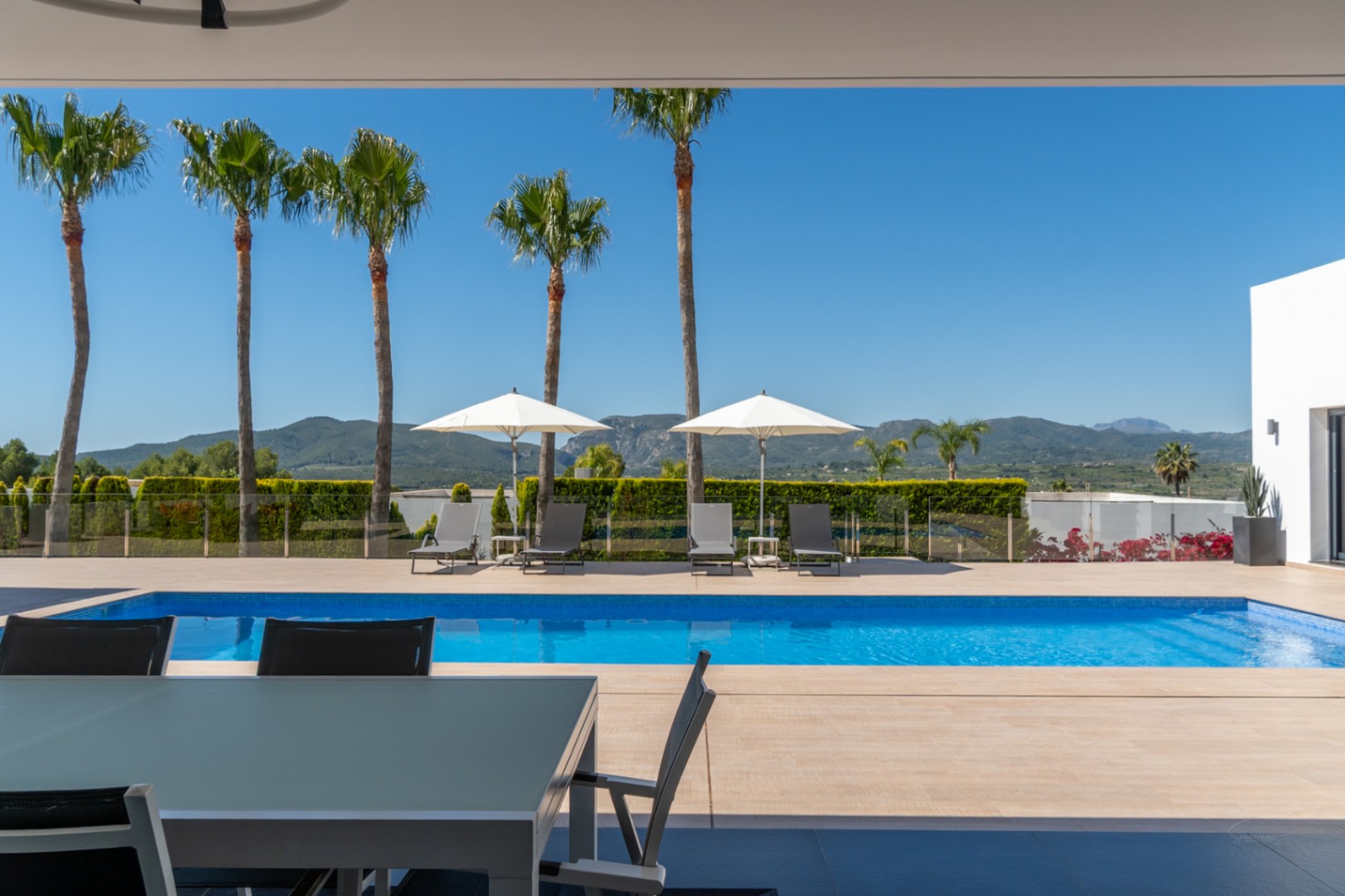 Terrasse couverte avec table à manger extérieure, offrant une vue sur la piscine carrelée de bleu, les palmiers et les montagnes lointaines.