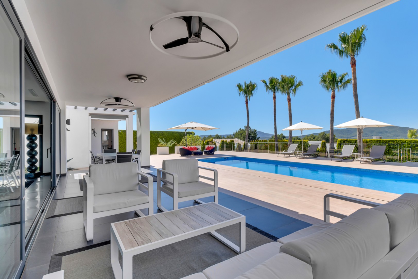 Terrasse couverte moderne avec mobilier de salon blanc, ventilateurs de plafond et grandes baies vitrées, s'ouvrant sur une piscine rectangulaire et des vues lointaines.