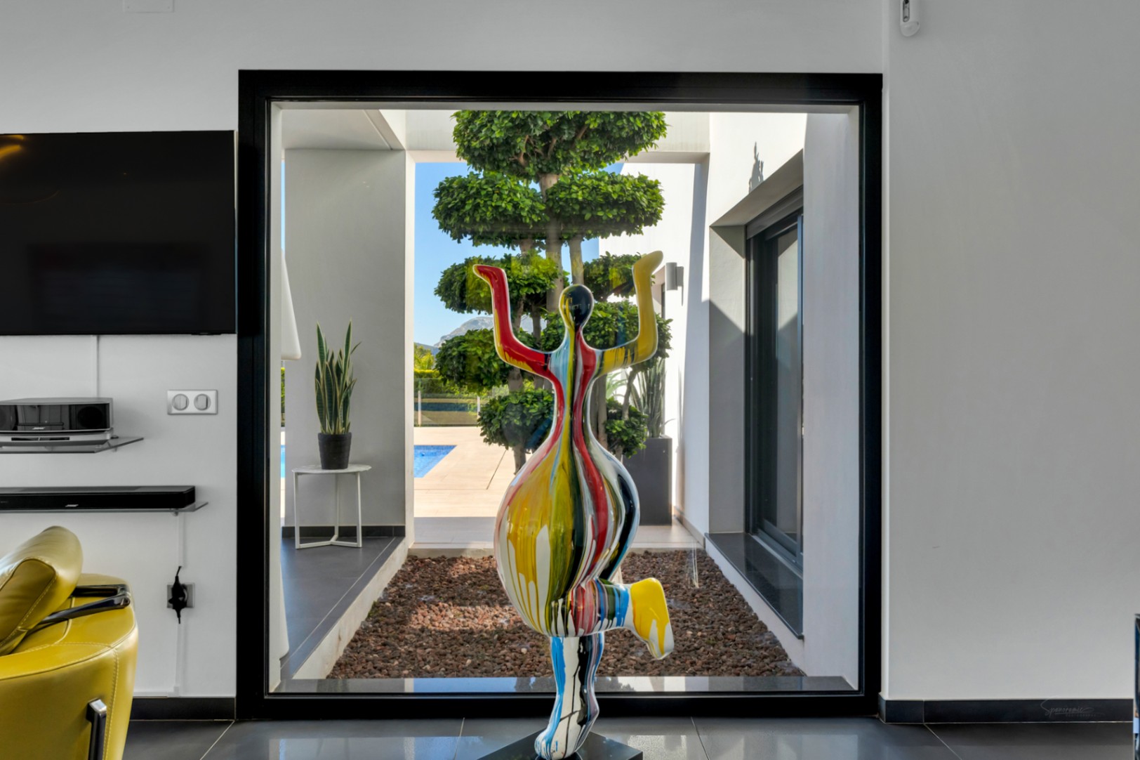 Une sculpture moderne et colorée encadre la vue depuis le salon à travers une grande fenêtre noire, donnant sur un jardin paysager avec un arbre unique et une piscine.