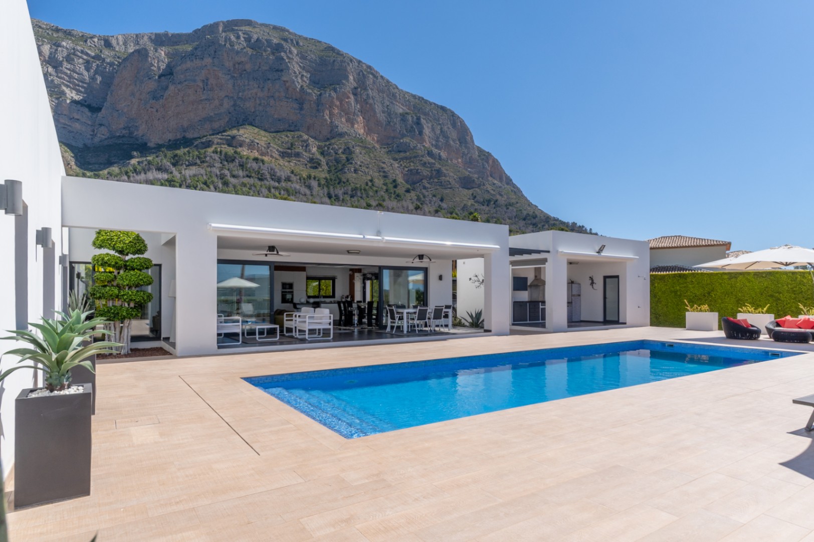 Vaste espace extérieur avec piscine rectangulaire en mosaïque bleue, terrasse carrelée claire et villa blanche moderne avec salon et salle à manger couverts intégrés, face au Montgó.