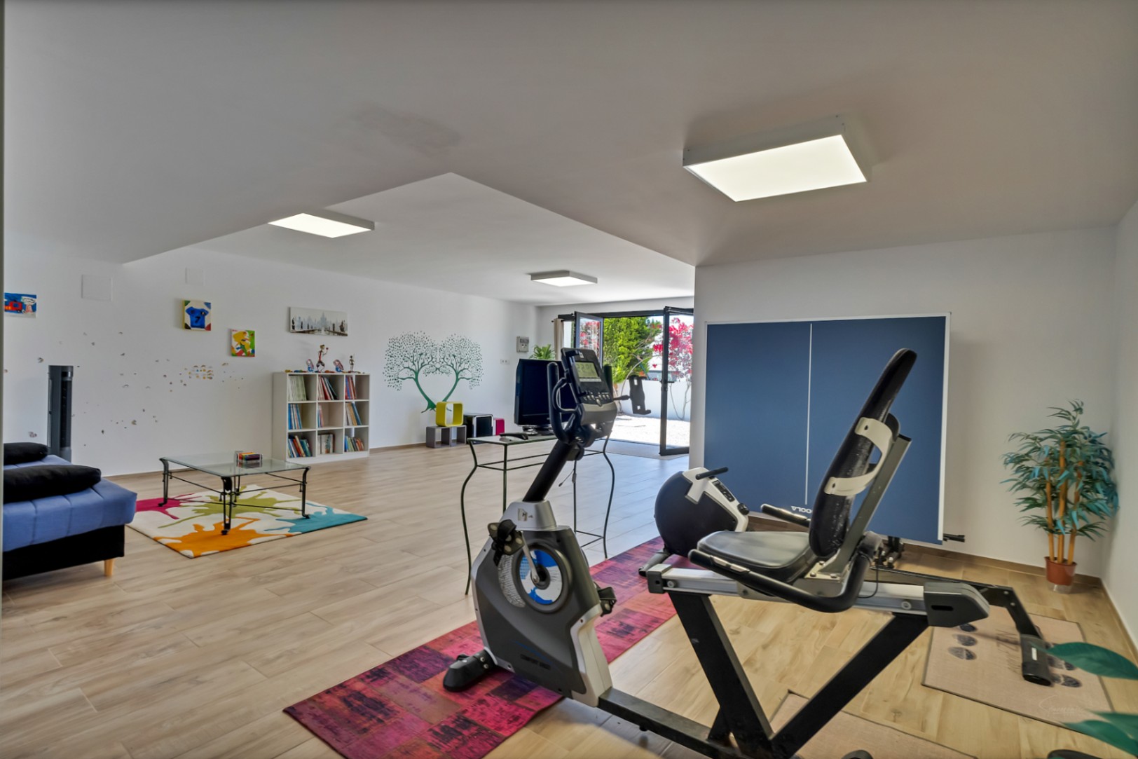 Vaste pièce polyvalente au niveau inférieur, équipée d'appareils de fitness, d'un coin salon avec canapé bleu et d'une bibliothèque. Accès direct à l'extérieur et parquet clair.