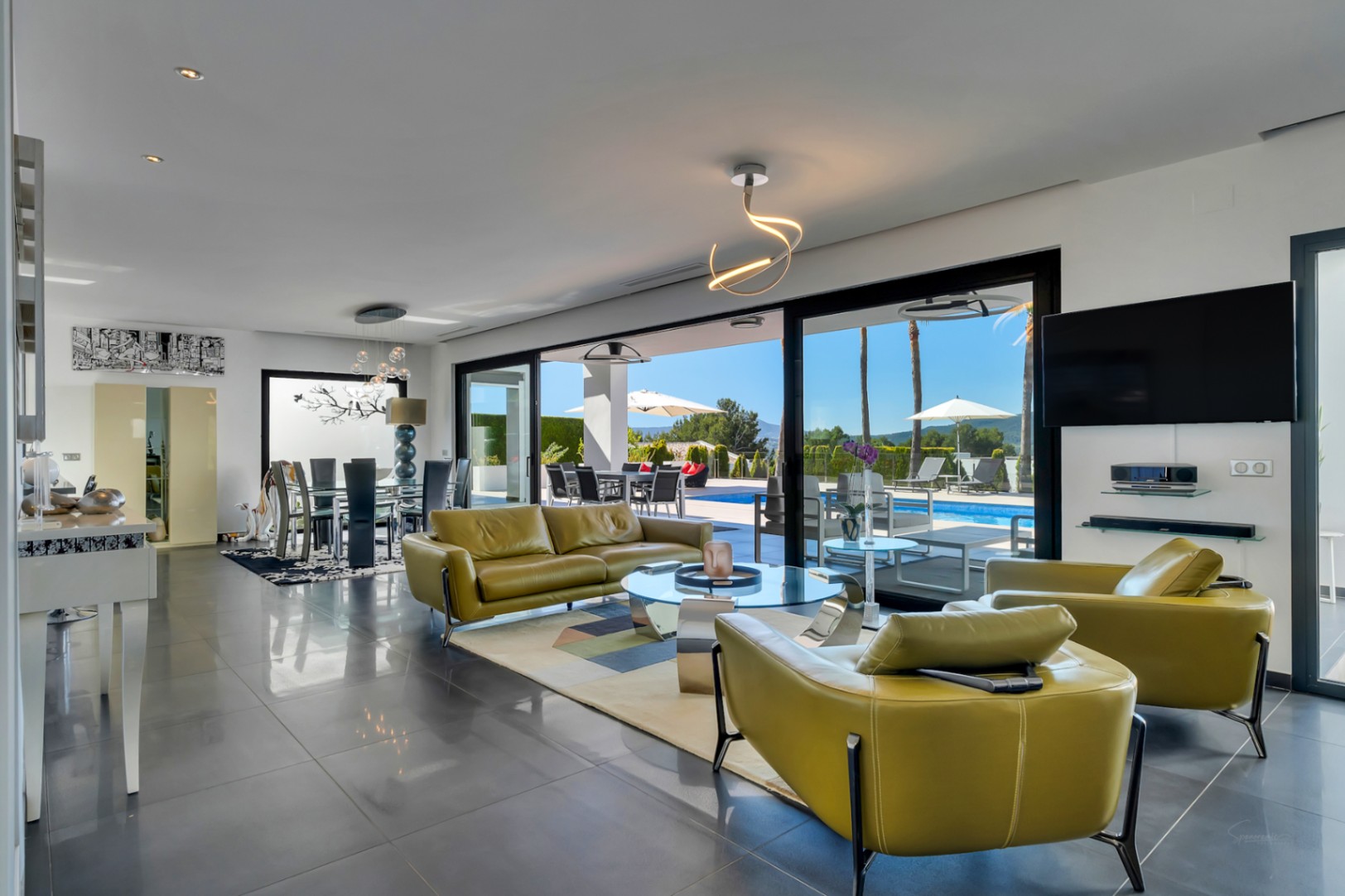 Vaste salon-salle à manger décloisonné avec accès direct à la terrasse et piscine chauffée via de grandes baies vitrées. Mobilier vert olive et sol carrelé gris foncé.