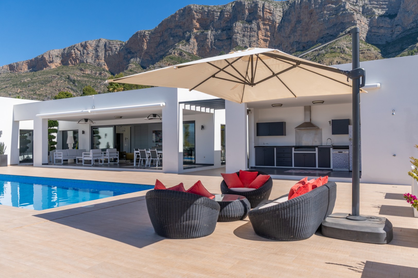 Vaste terrasse avec piscine, coin salon et cuisine extérieure équipée, encadrée par l'imposante montagne du Montgó sous un ciel bleu clair.