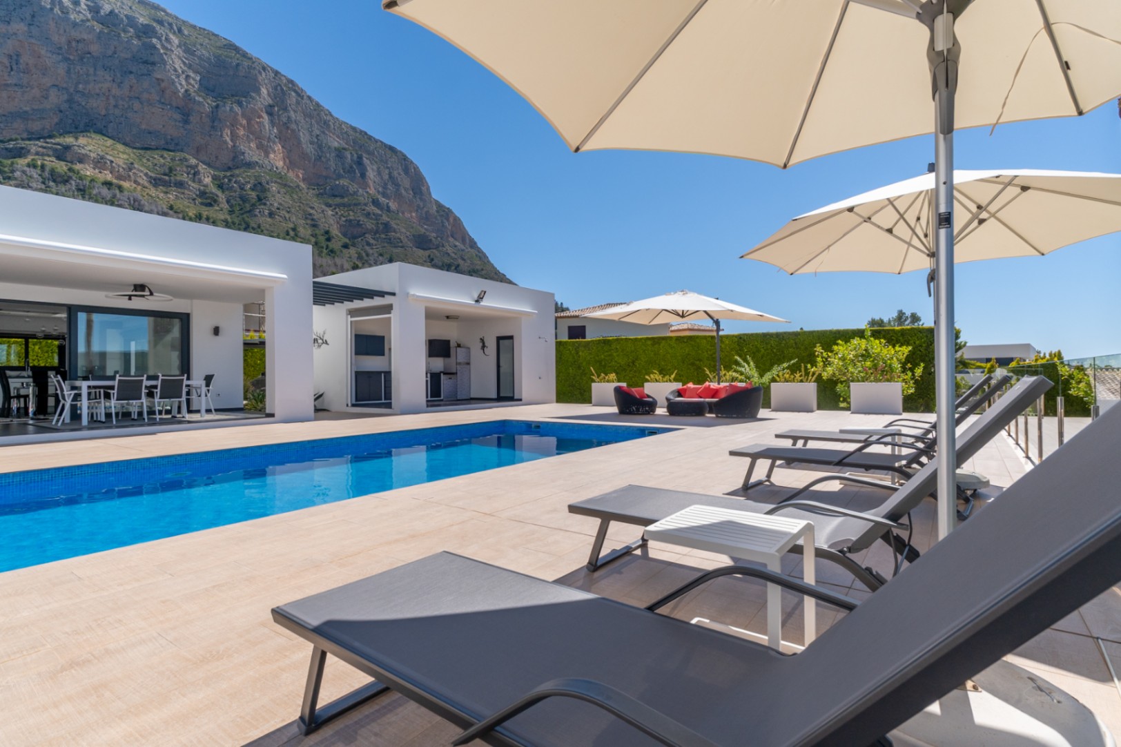 Vaste terrasse de piscine carrelée de bleu, espace barbecue extérieur et chaises longues sous parasols, avec vue sur l'imposant Montgó.