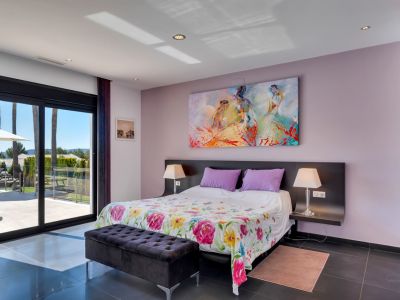 Chambre à coucher avec accès direct à la terrasse par de grandes portes coulissantes en verre, sol carrelé foncé, lit orné d'une couette florale et tableau abstrait coloré. Vues sur les palmiers extérieurs.