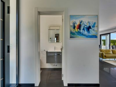 Couloir lumineux vers une salle de bain moderne avec meuble vasque suspendu, murs texturés et miroir éclairé. Un tableau abstrait et un aperçu du salon avec terrasse sont visibles.