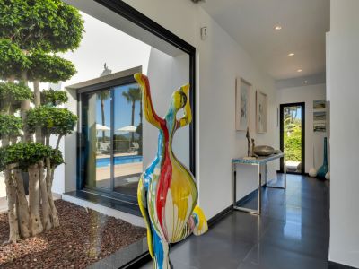 Couloir moderne aux carreaux sombres, orné d'une sculpture abstraite colorée et offrant une vue sur la terrasse piscine et un arbre décoratif intérieur.
