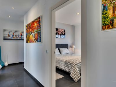 Couloir moderne avec carrelage gris foncé, éclairage encastré et art abstrait coloré, menant à une chambre avec lit à motifs et lampe de chevet.